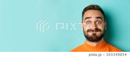 Headshot of funny bearded man showing faces and pouting, holding breath, standing in orange sweater against turquoise background 103349854