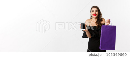 Beauty and shopping concept. Beautiful and stylish woman showing mobile phone screen and bag, buying online, standing over white background Beauty and shopping concept. Beautiful and stylish woman showing mobile phone screen and bag, buying online, standing over white background 103349860