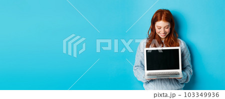 Cute redhead woman in sweater, showing and looking at laptop screen with pleased smile, demonstrating something online, standing over blue background Cute redhead woman in sweater, showing and looking at laptop screen with pleased smile, demonstrating something online, standing over blue background 103349936