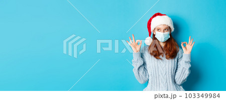 Christmas, quarantine and covid-19 concept. Cute teen redhead girl in santa hat and sweater, wearing face mask from coronavirus, showing okay signs, approve and praise something Christmas, quarantine and covid-19 concept. Cute teen redhead girl in santa hat and sweater, wearing face mask from coronavirus, showing okay signs, approve and praise something 103349984