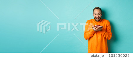 Handsome adult man celebrating birthday, blowing out candle on cake and making wish, standing against turquoise background Handsome adult man celebrating birthday, blowing out candle on cake and making wish, standing against turquoise background 103350023