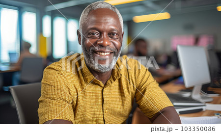 Portrait of a dark-skinned adult man with gray hair, who smiles and sits in the office at a laptop in a bright office, wearing a yellow shirt. AI generated. 103350086
