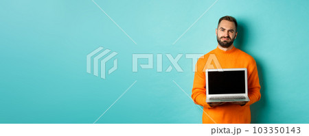 Handsome bearded man in orange sweater showing laptop screen, demonstrating promo, grimacing disappointed and upset standing over light blue background 103350143