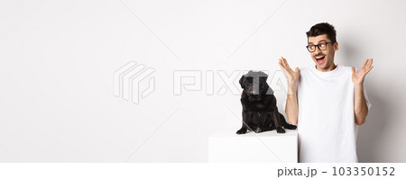 Image of cheerful young man rejoicing, looking at cute black dog pug and smiling, standing over white background 103350152