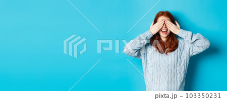 Cheerful redhead female model close eyes and waiting for christmas gift, holding hands on face and smiling amused, anticipating surprise, standing over blue background 103350231