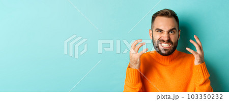 Close-up of angry caucasian man looking mad, grimacing and shaking hands, having break down, standing over light blue background Close-up of angry caucasian man looking mad, grimacing and shaking hands, having break down, standing over light blue background 103350232