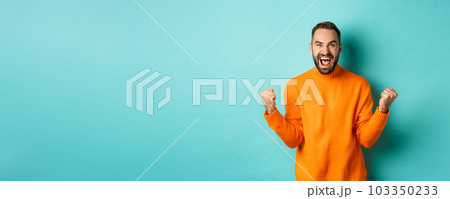 Excited man celebrating victory, rejoicing and making fist pump gesture, winning and looking satisfied, saying yes, achieve goal, standing over light blue background 103350233