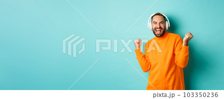 Happy adult man in orange sweater, looking up and listening music in headphones, standing over blue background Happy adult man in orange sweater, looking up and listening music in headphones, standing over blue background 103350236