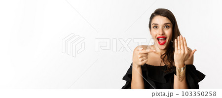 Happy young woman saying yes, become a bride, pointing at engagement ring on finger and smiling pleased, standing over white background 103350258