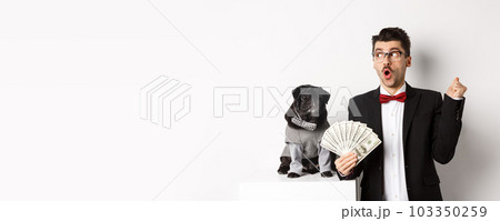 Happy young man in suit earn money with his dog. Guy rejoicing, holding dollars and staring left, black pug in costume staring at camera, white background 103350259
