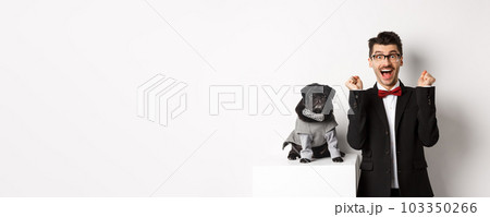Animals, party and celebration concept. Happy young man in suit and puppy in pet cosume standing over white background, dog owner rejoicing and triumphing Animals, party and celebration concept. Happy young man in suit and puppy in pet cosume standing over white background, dog owner rejoicing and triumphing 103350266
