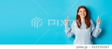 Winter holidays and people concept. Cute teenage redhead girl pointing fingers up, looking at top promo and smiling amused, standing over blue background Winter holidays and people concept. Cute teenage redhead girl pointing fingers up, looking at top promo and smiling amused, standing over blue background 103350267