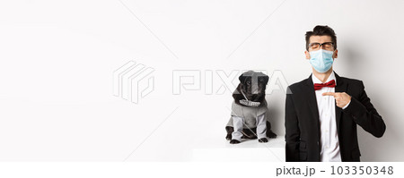 Coronavirus, pets and celebration concept. Disappointed young man in face mask and suit, pointing finger at cute black pug dog wearing party costume, standing over white background 103350348