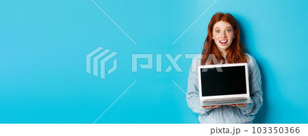Excited redhead female freelancer showing laptop screen, staring at camera amazed, standing with computer against blue background 103350366