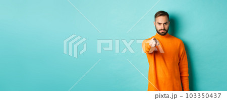 Confident man blame you, frowning and pointing finger at camera, accuse person, judgemental stare, standing over light blue background 103350437