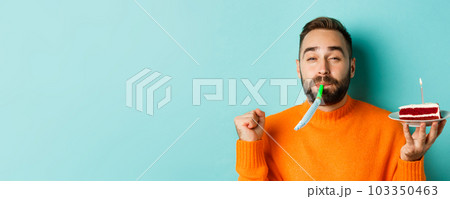 Close-up of funny adult man celebrating his birthday, holding bday cake with candle, blowing party wistle and rejoicing, standing over light blue background 103350463
