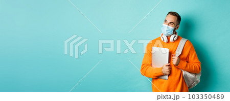 Handsome caucasian man with headphones and backpack, holding laptop and wearing medical mask, looking happy, standing over light blue background Handsome caucasian man with headphones and backpack, holding laptop and wearing medical mask, looking happy, standing over light blue background 103350489