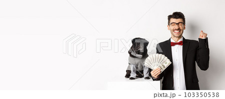 Happy young man and cute black dog standing in party costumes, pug owner holding money dollars and rejoicing, winning prize, white background Happy young man and cute black dog standing in party costumes, pug owner holding money dollars and rejoicing, winning prize, white background 103350538