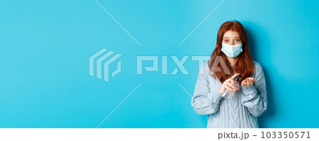 Winter, covid-19 and social distancing concept. Satisfied young redhead woman in face mask showing alright, okay gestures and looking left at promo, blue background 103350571