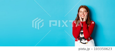 Winter holidays and Christmas Eve concept. Cheerful and cute redhead girl in xmas sweater, blushing and touching cheeks, smiling and rejoicing over blue background 103350623