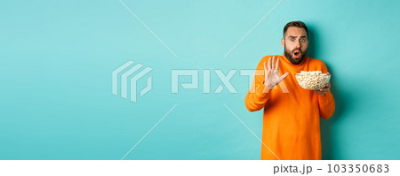 Portrait of scared guy holding bowl of popcorn and declining something, showing stop gesture, standing over blue background 103350683