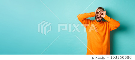 Image of funny and happy guy holding hands on eyes, looking through fake glasses, smiling amused, standing in orange sweater against turquoise background 103350686