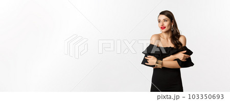 Beauty and fashion concept. Elegant and beautiful woman in black dress, makeup, embracing herself and looking away with sensual gaze, standing over white background 103350693