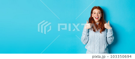 Supportive redhead girl in sweater, showing thumbs-up and looking amazed, praising good choice, standing over blue background Supportive redhead girl in sweater, showing thumbs-up and looking amazed, praising good choice, standing over blue background 103350694