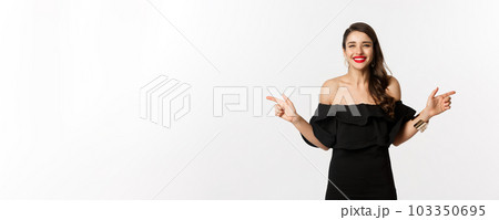 Fashion and beauty. Attractive woman in jewelry, makeup and black dress, smiling and pointing fingers sideways copy space offer, white background 103350695