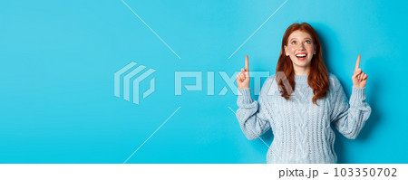 Winter holidays and people concept. Beautiful teenage girl with red hair, wearing sweater, pointing fingers up at logo and smiling, showing advertisement, blue background 103350702
