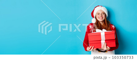 Happy holidays and Christmas concept. Cheerful teen girl with red hair, wearing santa hat and holding xmas presents, 103350733