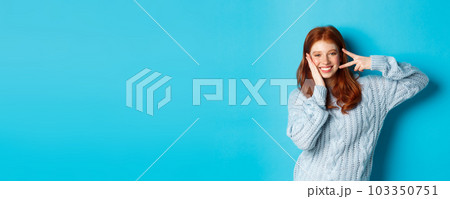 Cheerful redhead female model sending good vibes, smiling and showing peace sign, standing over blue background 103350751