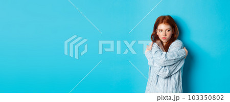 Silly and cute redhead girl pucker lips and looking offended, hugging herself and looking away offended, sulking and being upset, standing over blue background 103350802