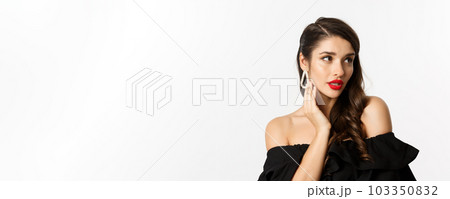 Fashion and beauty concept. Close-up of elegant woman in black dress, showing earrings and looking sensual, red lipstick and makeup on, white background 103350832