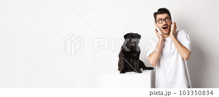 Amazed young man standing with cute black puppy, staring at upper right corner surprised and excited, standing near pug over white background Amazed young man standing with cute black puppy, staring at upper right corner surprised and excited, standing near pug over white background 103350834