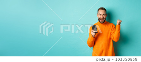 Photo of happy man triumphing, holding mobile phone and make winner gesture, rejoice and achieve goal, standing over light blue background Photo of happy man triumphing, holding mobile phone and make winner gesture, rejoice and achieve goal, standing over light blue background 103350859
