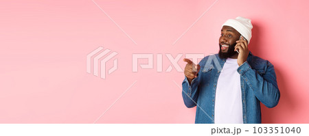 Handsome modern Black man talking on mobile phone, pointing left at person and smiling, standing over pink background Handsome modern Black man talking on mobile phone, pointing left at person and smiling, standing over pink background 103351040