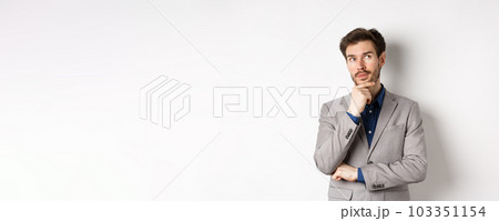 Pensive handsome man in suit looking up and thinking, making choice, pondering ideas, standing against white background Pensive handsome man in suit looking up and thinking, making choice, pondering ideas, standing against white background 103351154