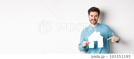Real estate and insurance concept. Smiling young man pointing at paper house cutout and looking happy, buy apartment, standing over white background 103351195