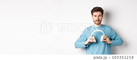 Young bearded guy in blue sweatshirt put on wireless headphones, listening music, standing on white background 103351219