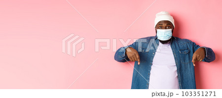 Covid-19, lifestyle and lockdown concept. Handsome african-american man in face mask, pointing fingers down, showing promo offer on bottom, standing over pink background Covid-19, lifestyle and lockdown concept. Handsome african-american man in face mask, pointing fingers down, showing promo offer on bottom, standing over pink background 103351271