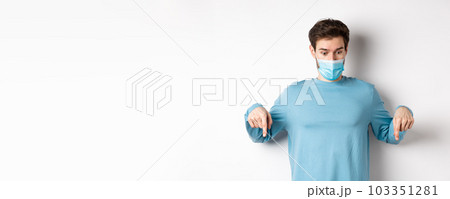 Covid-19, health and quarantine concept. Curious man checking out advertisement, pointing and looking down, wearing medical mask with casual blue sweatshirt, white background Covid-19, health and quarantine concept. Curious man checking out advertisement, pointing and looking down, wearing medical mask with casual blue sweatshirt, white background 103351281