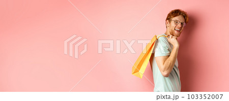 Carefree young man with red hair and glasses, walking with shopping bag over his shoulder and smiling, shopper buying presents, standing satisfied over pink background Carefree young man with red hair and glasses, walking with shopping bag over his shoulder and smiling, shopper buying presents, standing satisfied over pink background 103352007
