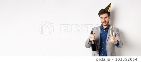 Celebration and holidays concept. Handsome man in suit and birthday hat giving glass of champagne, holding bottle alcohol, standing on white background Celebration and holidays concept. Handsome man in suit and birthday hat giving glass of champagne, holding bottle alcohol, standing on white background 103352054