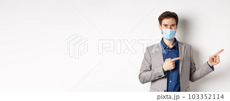 Covid-19, pandemic and business concept. Confident male manager in suit and medical mask showing way, pointing fingers right at advertisement, white background Covid-19, pandemic and business concept. Confident male manager in suit and medical mask showing way, pointing fingers right at advertisement, white background 103352114