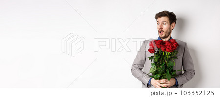 Handsome young man in suit holding red roses, looking left with surprised and startled expression, standing on Valentines day over white background Handsome young man in suit holding red roses, looking left with surprised and startled expression, standing on Valentines day over white background 103352125