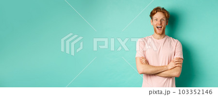 Enthusiastic redhead man in t-shirt checking out interesting promo, cross arms on chest and looking at camera with awe, standing over turquoise background 103352146