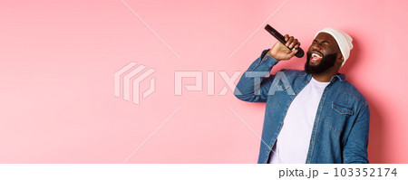 Handsome Black man in beanie and denim shirt singing karaoke, holding microphone, standing over pink background 103352174