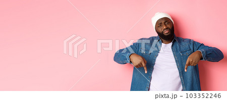 Skeptical and doubtful Black guy staring at camera unamused, pointing fingers down at something bad, standing over pink background 103352246