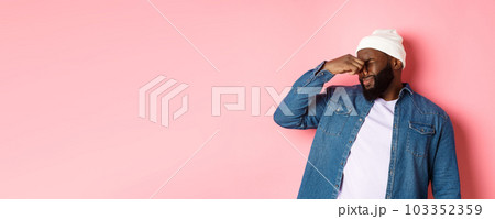 Disgusted african-american man shut nose from aversion, displeased by awful bad smell, standing in beanie and denim shirt over pink background 103352359